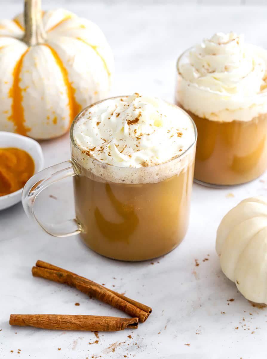 Pumpkin spice latte in glass mugs with whipped cream on top and cinnamon sticks and a white pumpkin in front of it.