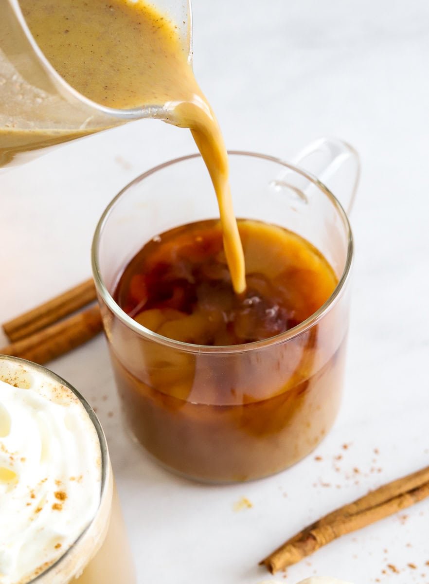 Pouring pumpkin coffee creamer into glass mug of coffee.