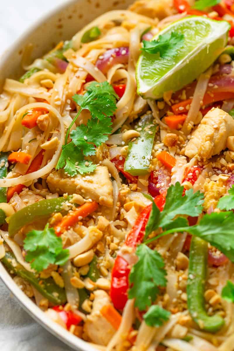 Cooked chicken and veggie pad thai in a pan topped with cilantro, peanuts and lime wedges.