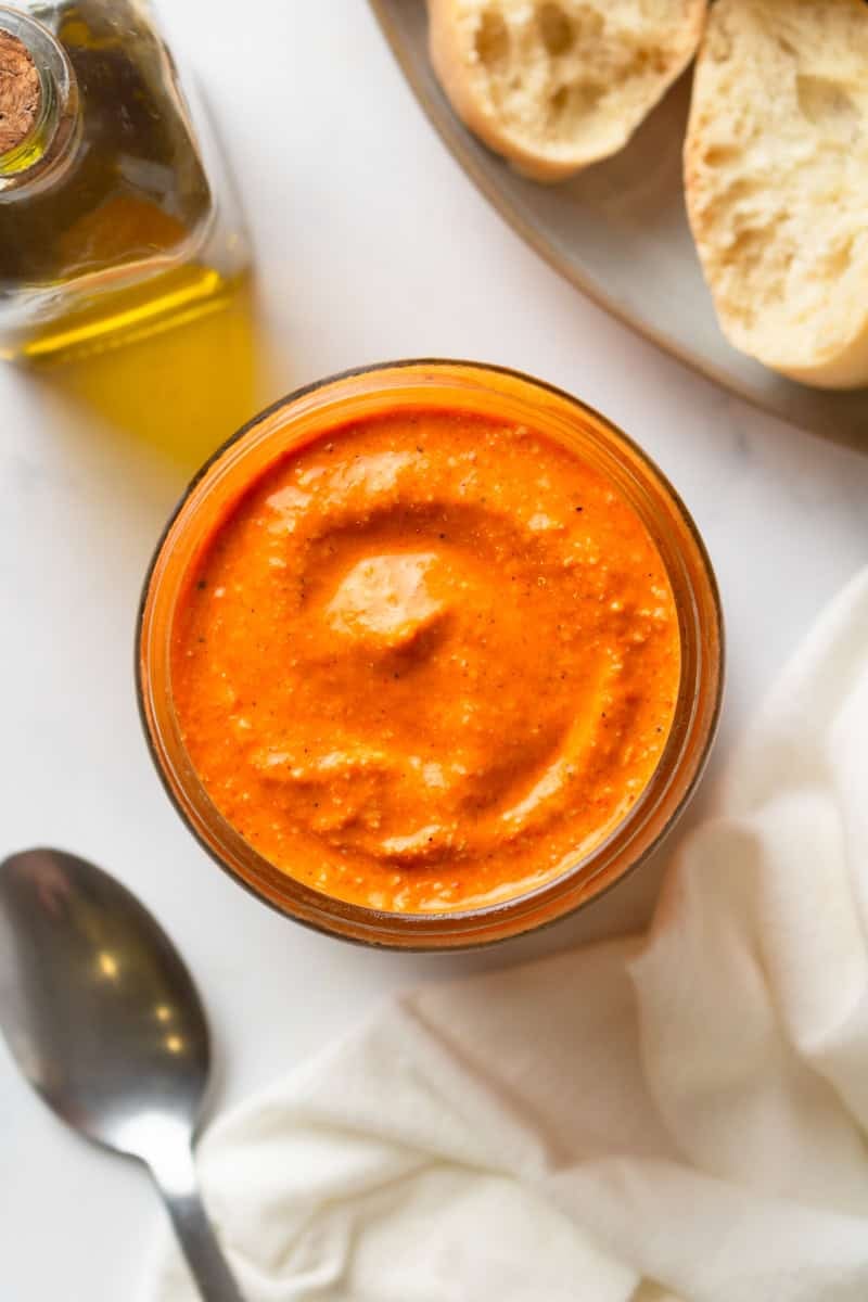 Romesco sauce in a glass jar with some bread and olive oil behind it and a spoon and linen in front of it. 