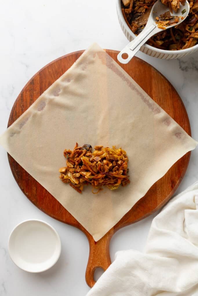 Egg roll wrapper on round wood board with egg rolls filling on it with more filling in a bowl behind it.
