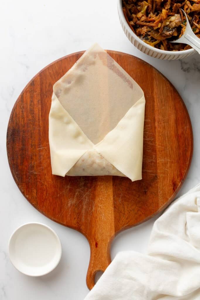 Round wood board with egg roll half wrapped on it with more egg roll filling in a bowl behind it.
