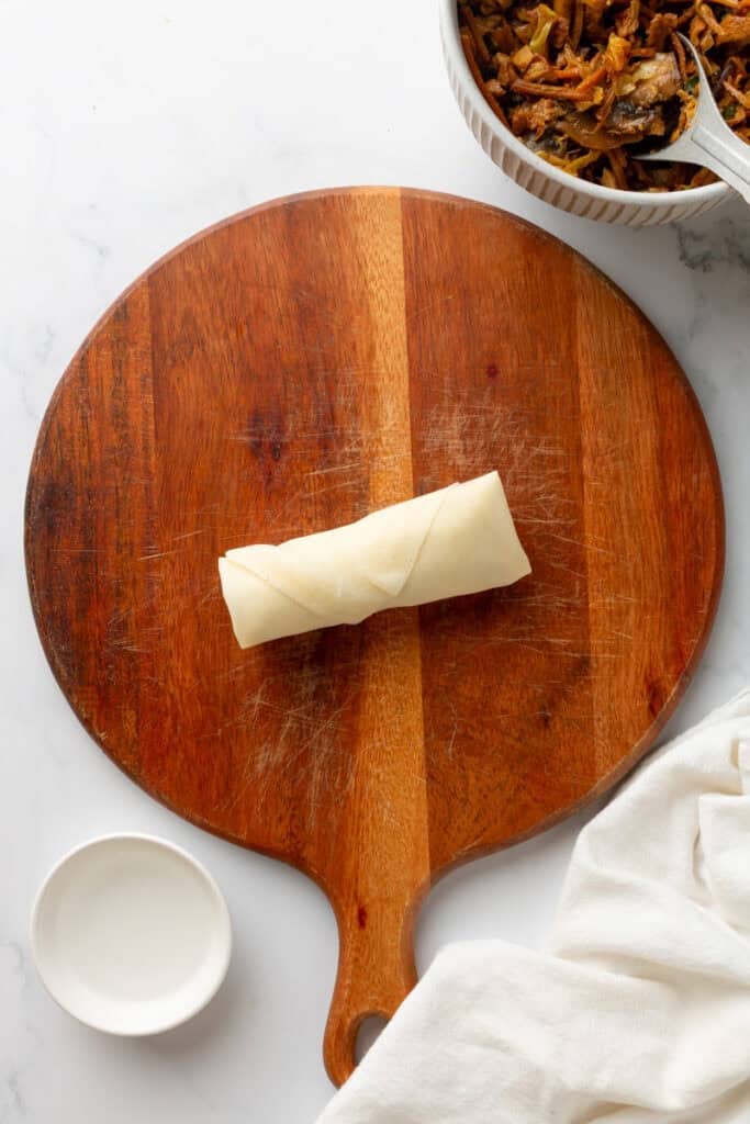 Round wooden board with un uncooked, rolled egg roll on it with filling in a bowl behind it and a white linen in front of it.