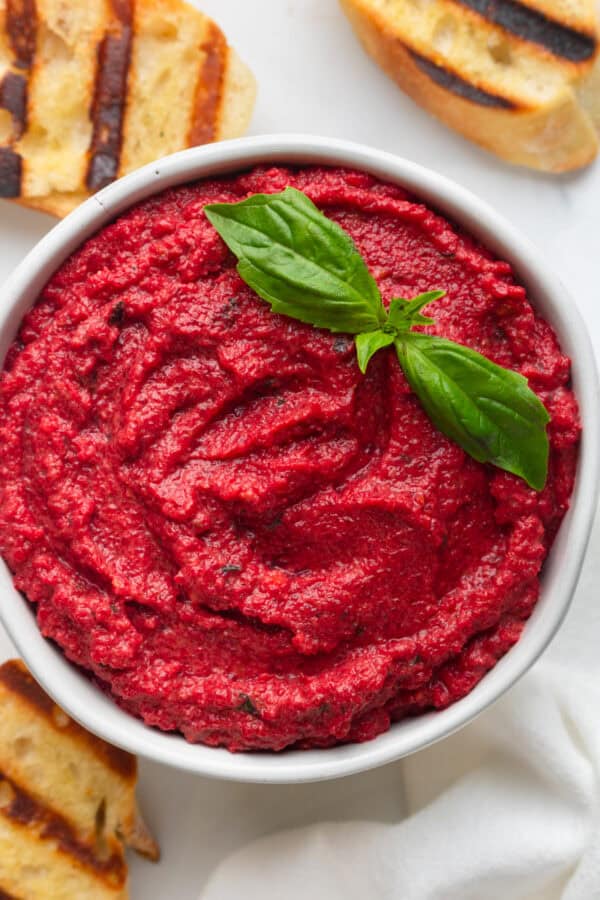 Beet pesto in a bowl with a piece of basil leaf on it with grilled bread around it.