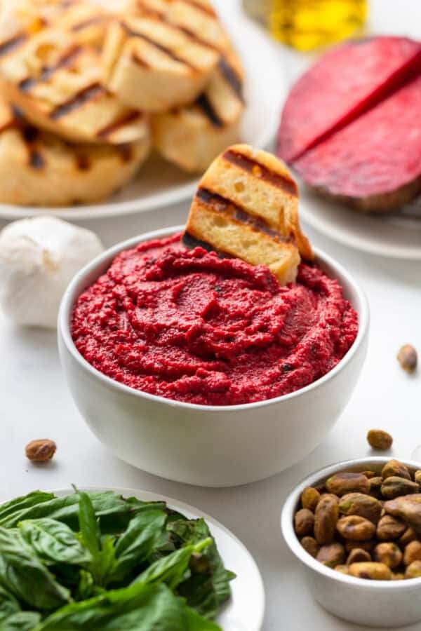 Bowl of pink beet pesto sauce with grilled bread slice in it with basil and pistachios in front of it and more grilled bread behind it.