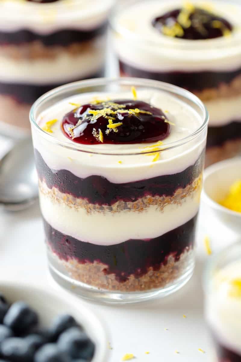 Layered blueberry cheesecake parfait with lemon zest on top in a clear glass with more parfaits behind it. 