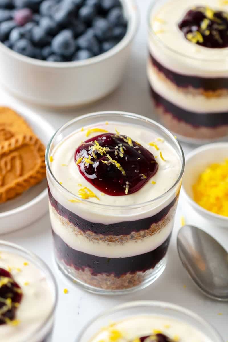Layered yogurt and blueberry parfait in a clear glass topped with pie filling and lemon zest with more behind it. 