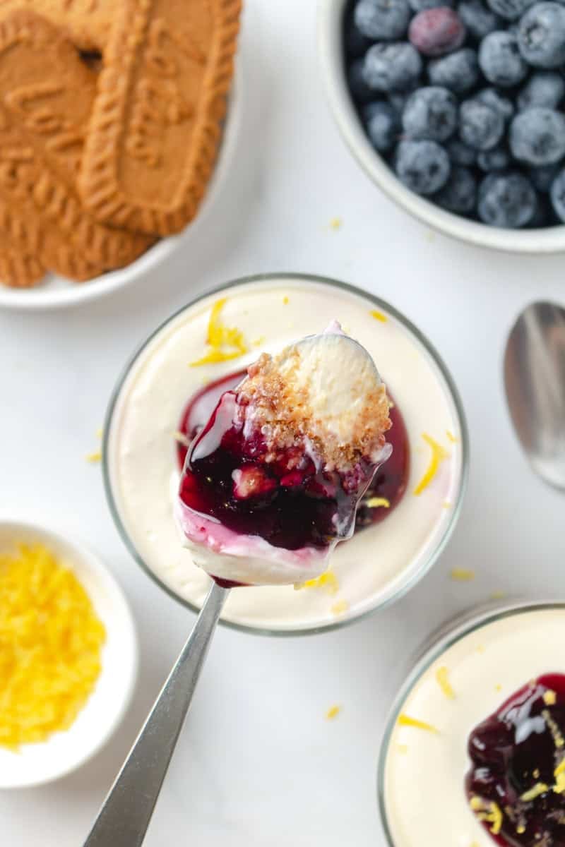 Spoon lifting filling from a cheesecake blueberry parfait in a glass with cookies and a bowl of blueberries behind it and lemon zest and another parfait in front of it. 