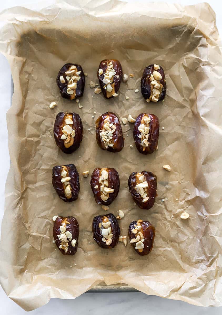 Sliced dates filled with peanut butter and chopped peanuts in rows on a lined baking sheet.