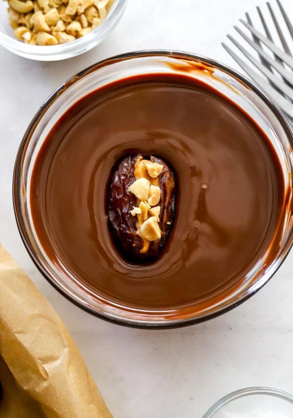 Date snickers in bowl of melted chocolate with more ingredients around it.