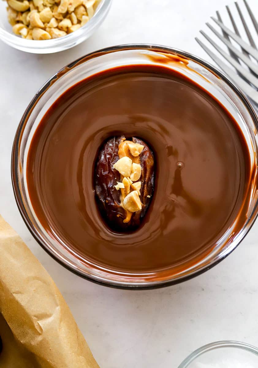 Date snickers in bowl of melted chocolate with more ingredients around it.