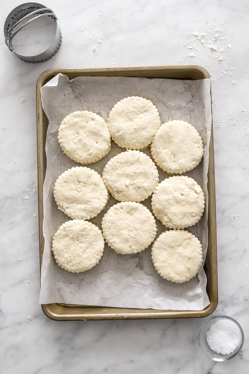 Cut out uncooked biscuits on a lined pan with salt in a bowl in front of it and a biscuit cutter behind it.