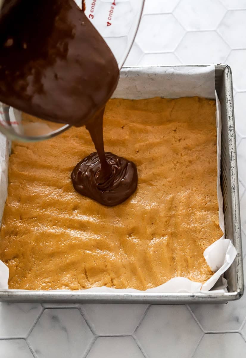 Pouring melted chocolate into a pan filled with peanut butter bar base in a metal lined pan.