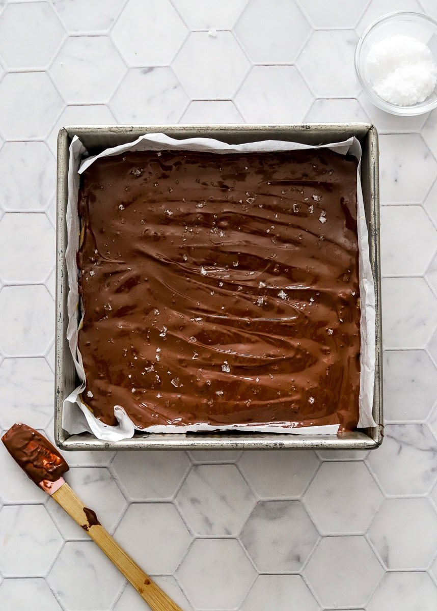Square metal pan filled with chocolate covered layered bars with a spatula with a wood handle with chocolate on it next to it.