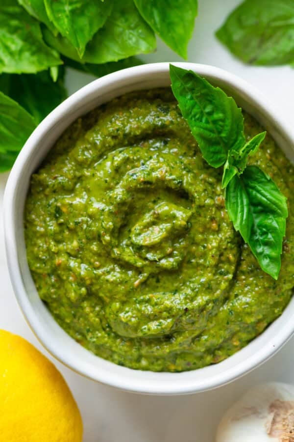 Round bowl filled with homemade pesto recipe with basil leaf on it with more basil behind it.