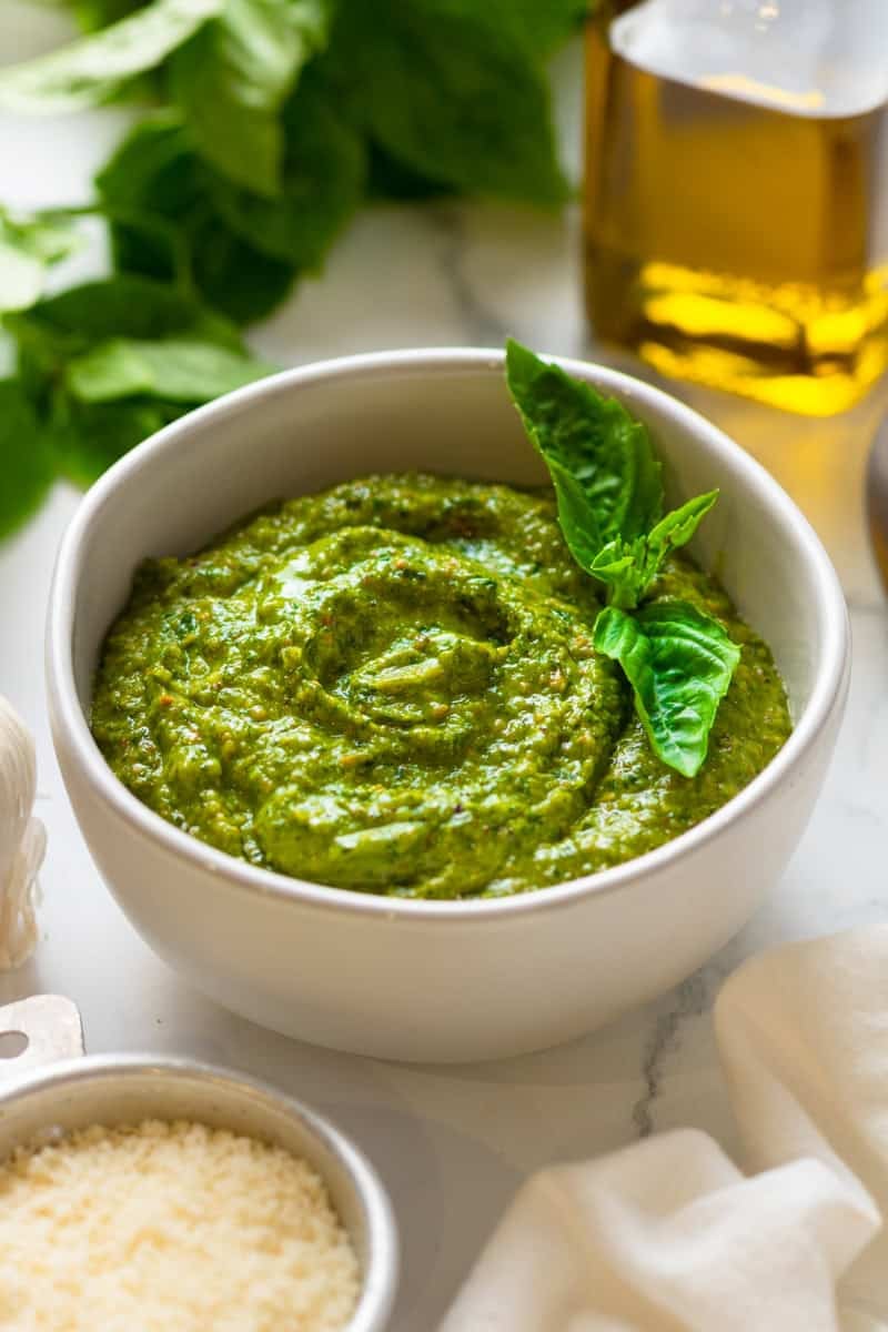 Green basil pesto in a bowl with basil leaves and olive oil behind it.