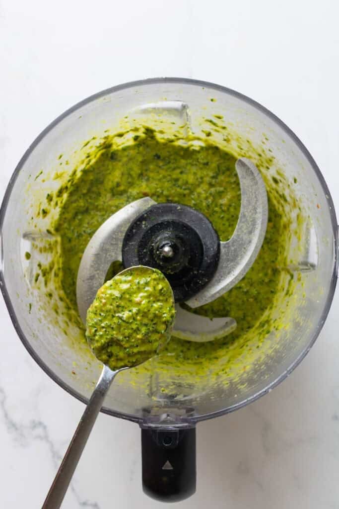 Blended homemade pesto in a food processor with a spoon with some not it over it.