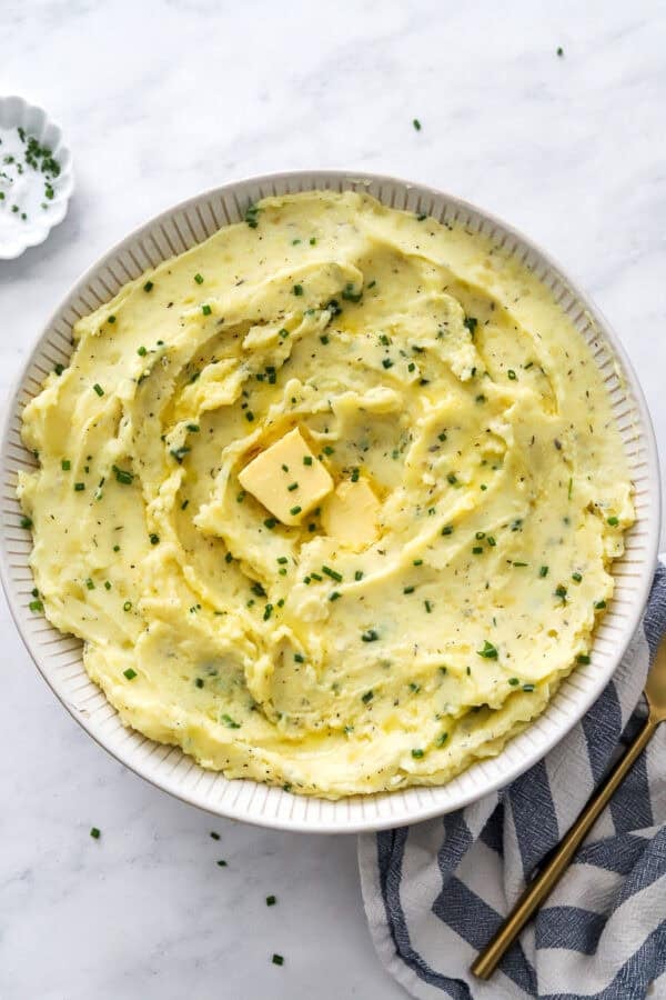 Bowl of healthy herby mashed potatoes with butter on it and a gold spoon on a striped towel next to it.