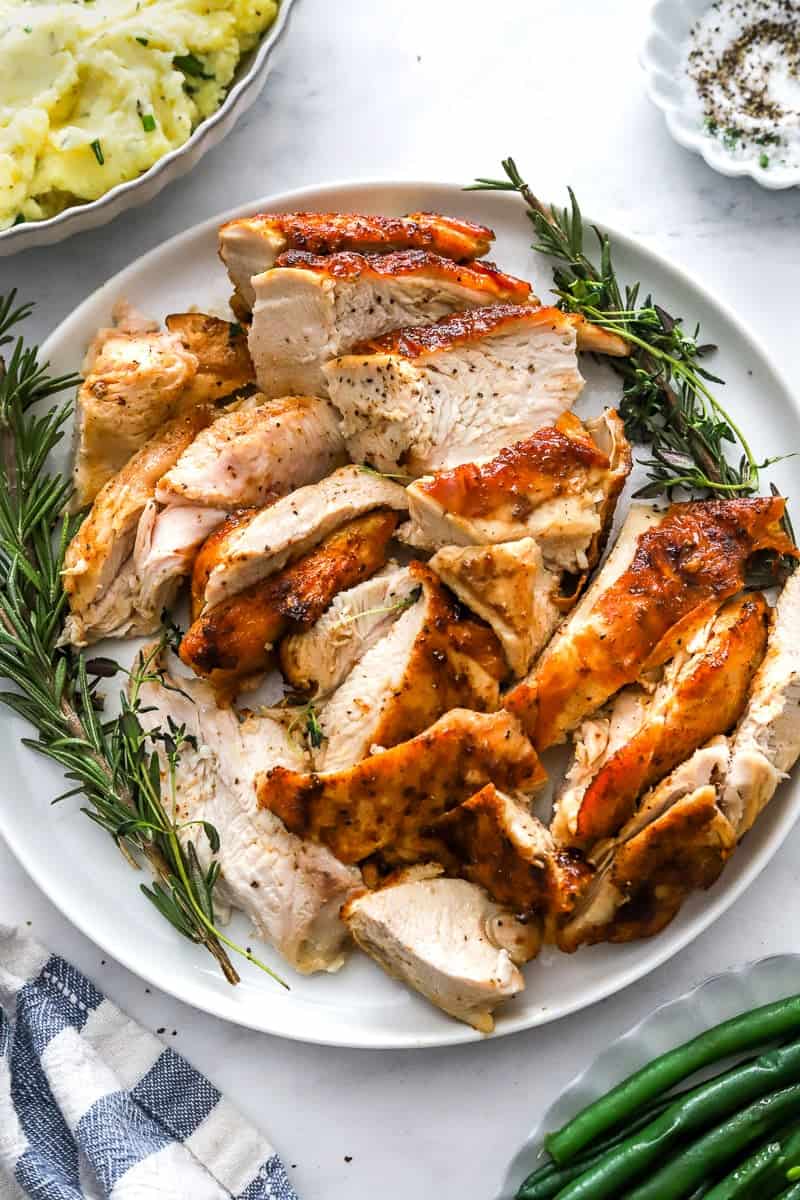Sliced, cooked turkey breast on a plate with rosemary around it with green beans in front of it.