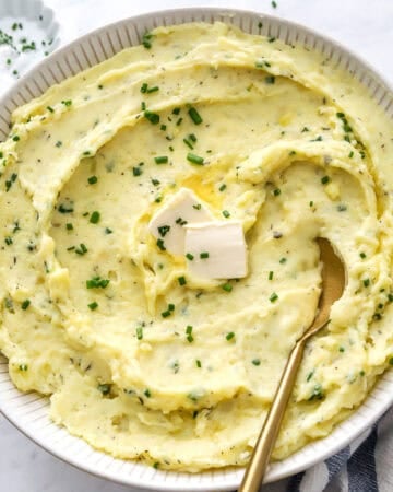 Round bowl filled with healthy mashed potatoes topped with herbs and butter with a gold serving spoon in the bowl.