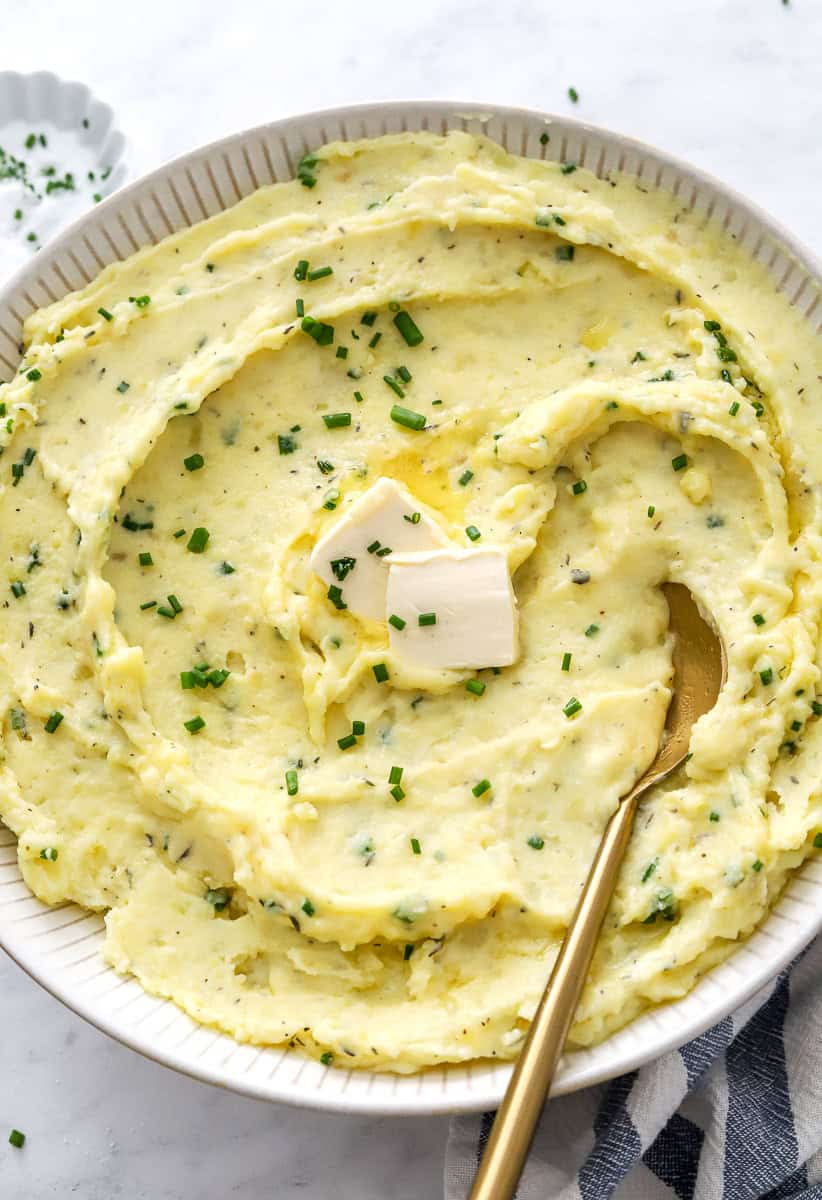 Round bowl filled with healthy mashed potatoes topped with herbs and butter with a gold serving spoon in the bowl.