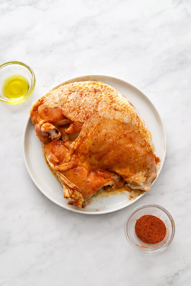 Uncooked turkey breast seasoned on a plate with bowl of seasoning in front of it and bowl of olive oil behind it.