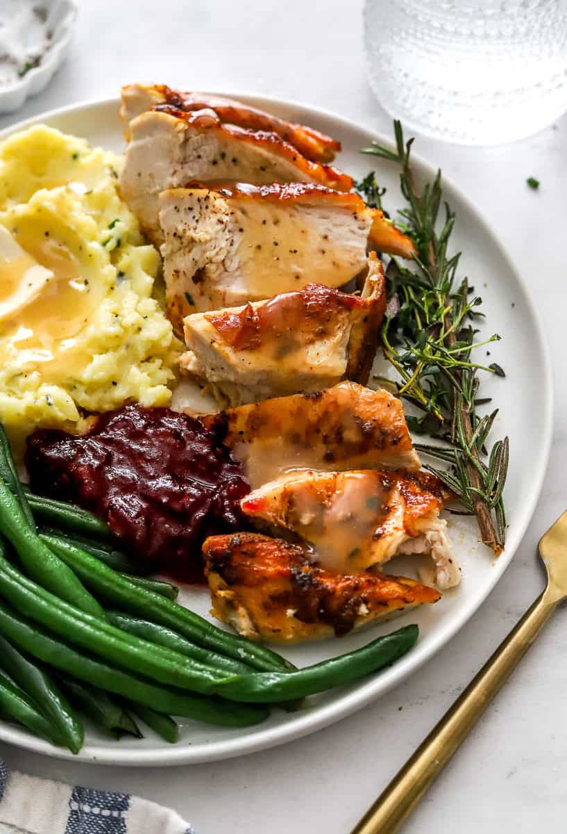 Sliced cooked turkey breast on a plate with mashed potatoes, green beans and cranberry sauce.