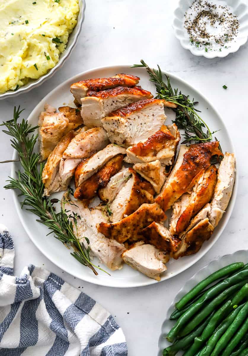 Sliced turkey on a plate with herbs around it with green beans in front of it and mashed potatoes in a bowl behind it.