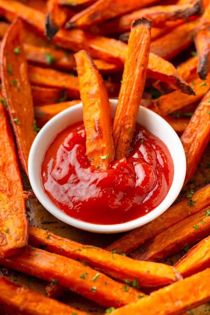 Baked sweet potato fries with some dipped in a small bowl of ketchup with more around it. 