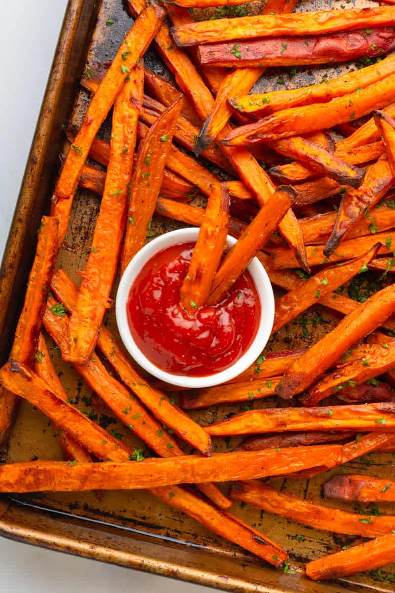 Baking pan with oven bake sweet potato fries with a small bowl o ketchup with a couple fries in it in the middle of the pan. 