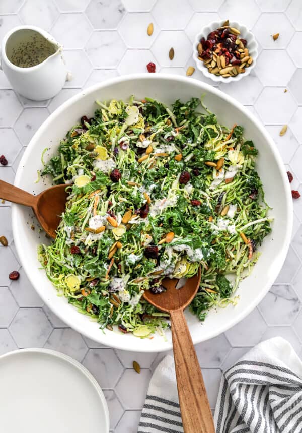 Mixed and dressed Brussels slaw salad with kale in a white bowl with serving spoons in the bowl and plates and a linen in front of it.