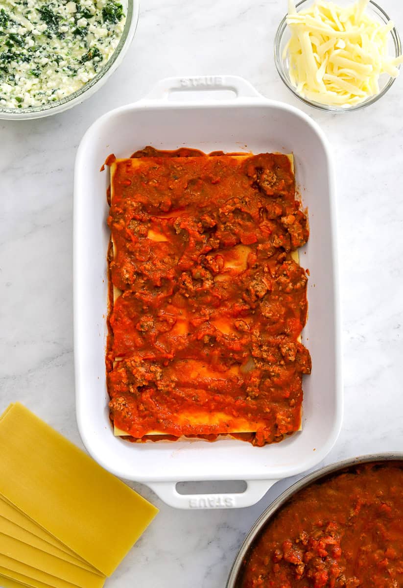 White lasagna pan filled with layered lasagna noodles, and sauce with the ingredients in bowls around it.