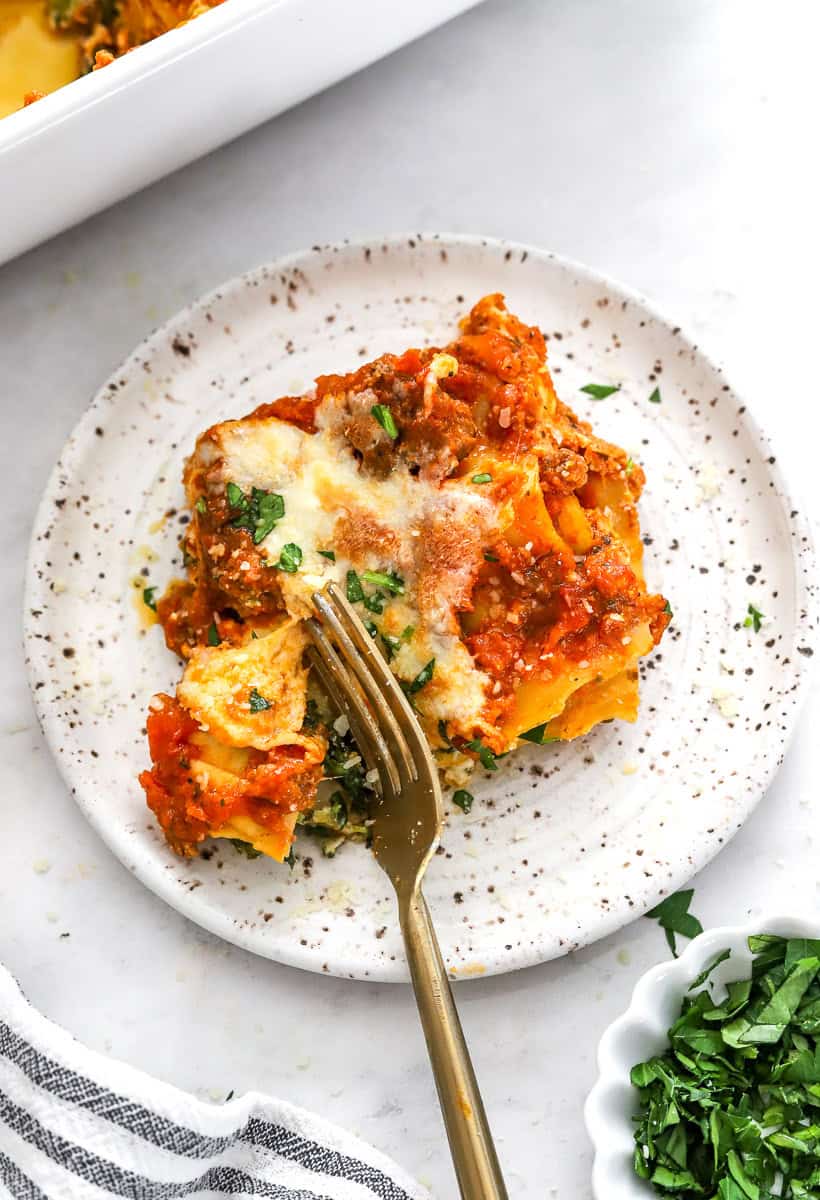Plate of slice of lasagna with a fork cutting through it with more lasagna in a pan behind it.