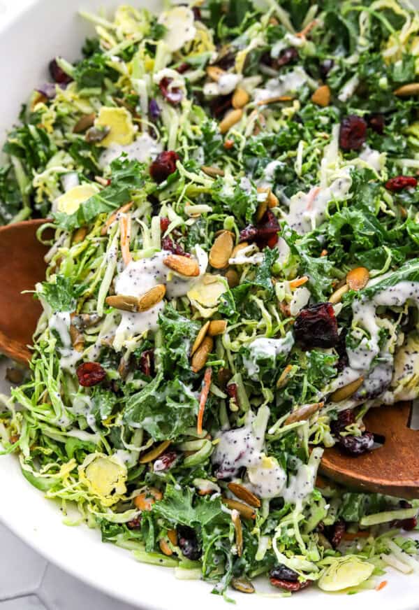Kale salad with Brussels sprouts and poppyseeds dressing mixed in a salad bowl with wood serving spoons in the bowl with it.