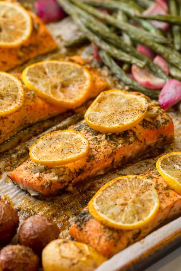 Roasted herb salmon on a baking pan with slices of lemon in top of it with green beans and potatoes around it.