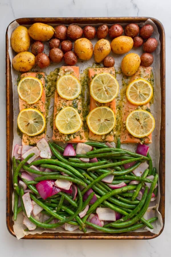 Cooked veggies and uncooked marinated salmon on a baking sheet with lemon slices on top of the salmon.