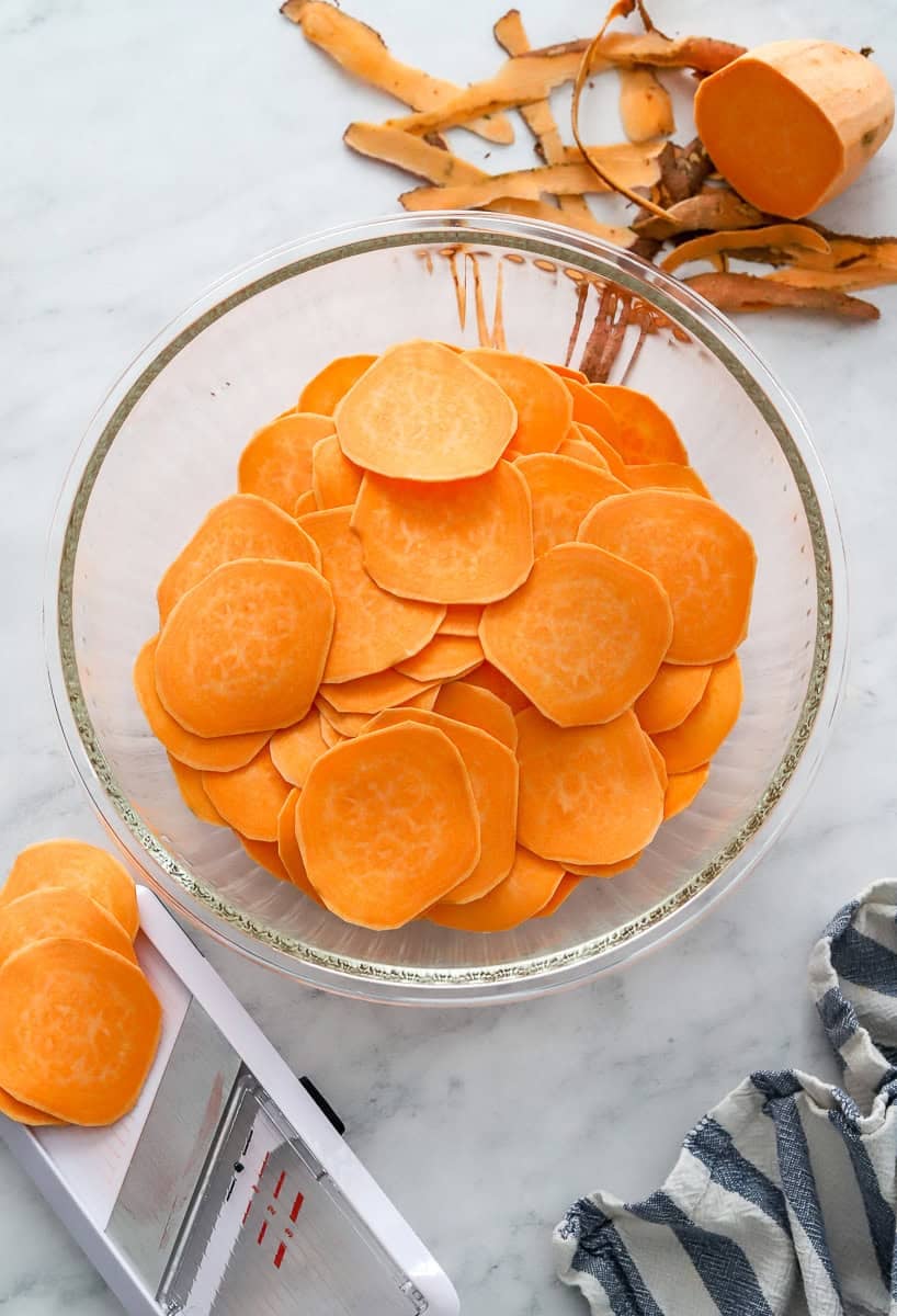 Sliced sweet potatoes in a bowl with a mandolin slicer in front of it.