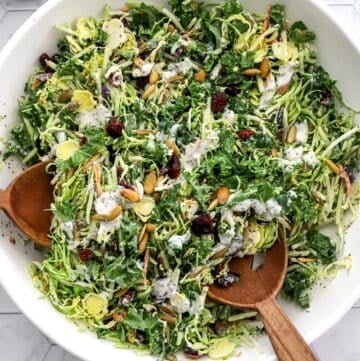 Sweet kale salad mixed with poppyseed dressing in a white salad bowl with wood serving spoons in the bowl with it.
