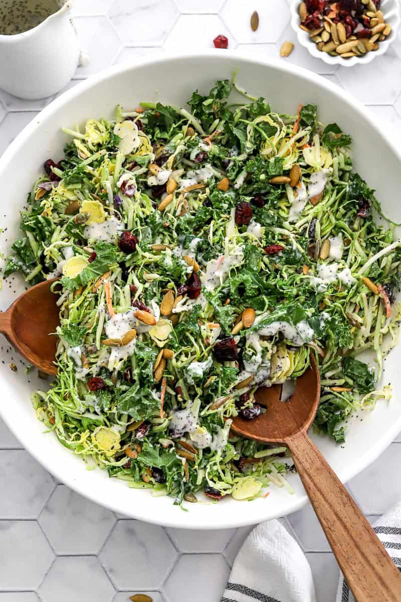 Sweet kale salad mixed with poppyseed dressing in a white salad bowl with wood serving spoons in the bowl with it.