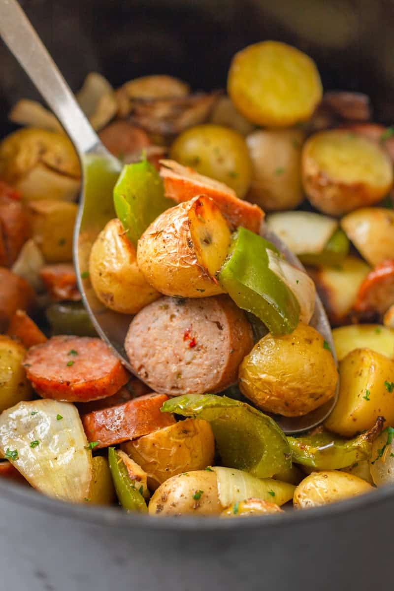Silver serving spoon with cooked cut sausage and potatoes and bell peppers on it with more in the air fryer behind it. 