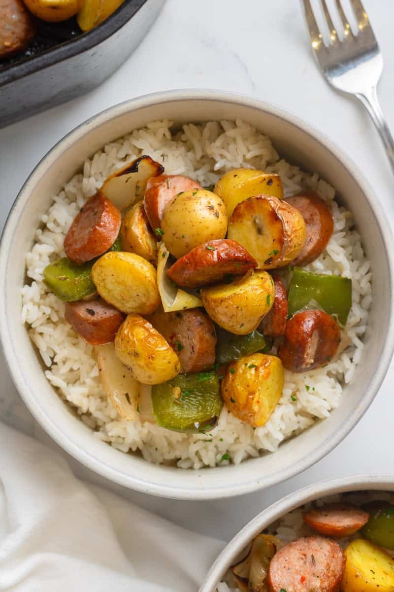 Bowl filled with rice with air fired potatoes and sausage slices with bell peppers with another bowl of it in front of it. 