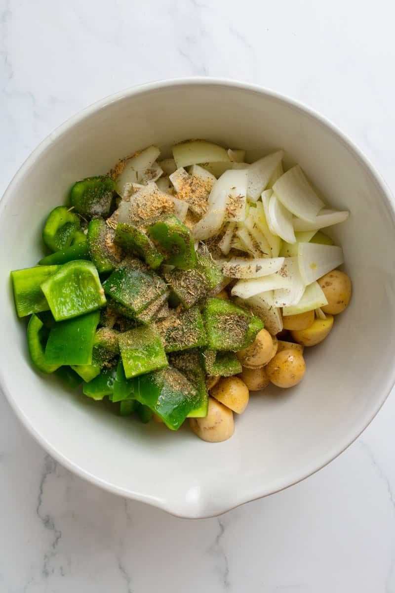 Chopped green peppers, chopped onion and cut gold potatoes with seasoning on it in a mixing bowl. 