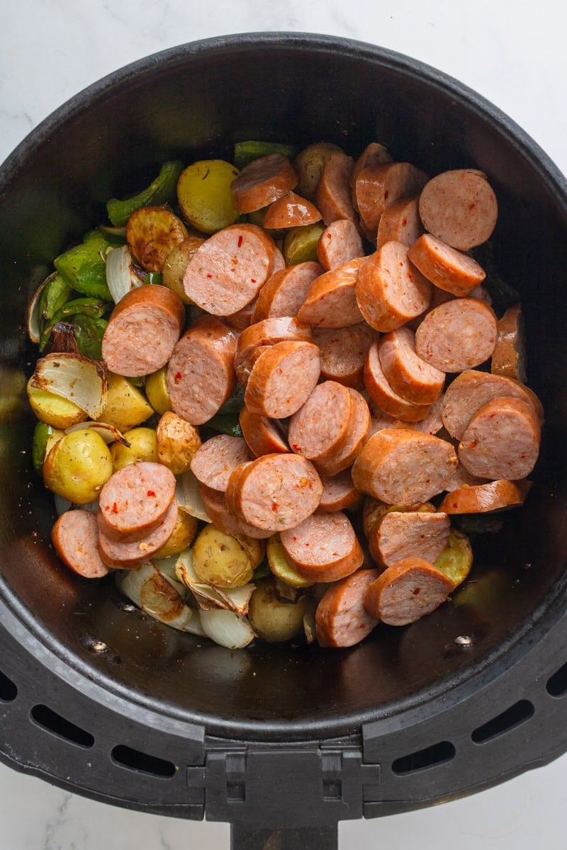 Cooked potatoes and peppers with uncooked sausage slices on top of it in a black air fryer basket. 
