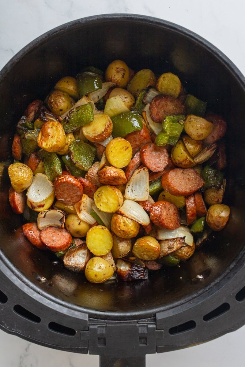 Cooked sausage and potatoes with peppers and onion in a round black air fryer basket. 