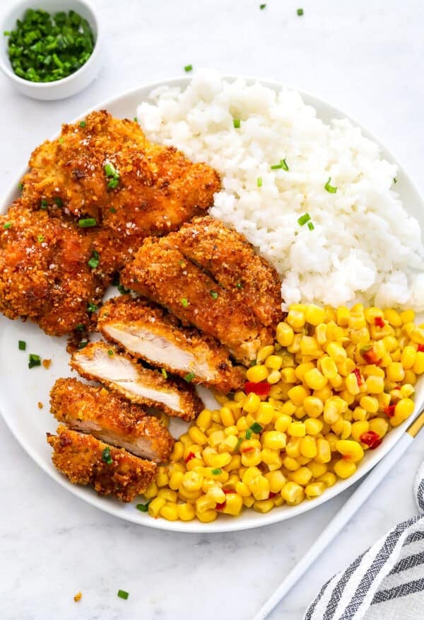 Air fried breaded chicken thighs on a white plate with rice and corn with one of the pieces of chicken cut into strips.