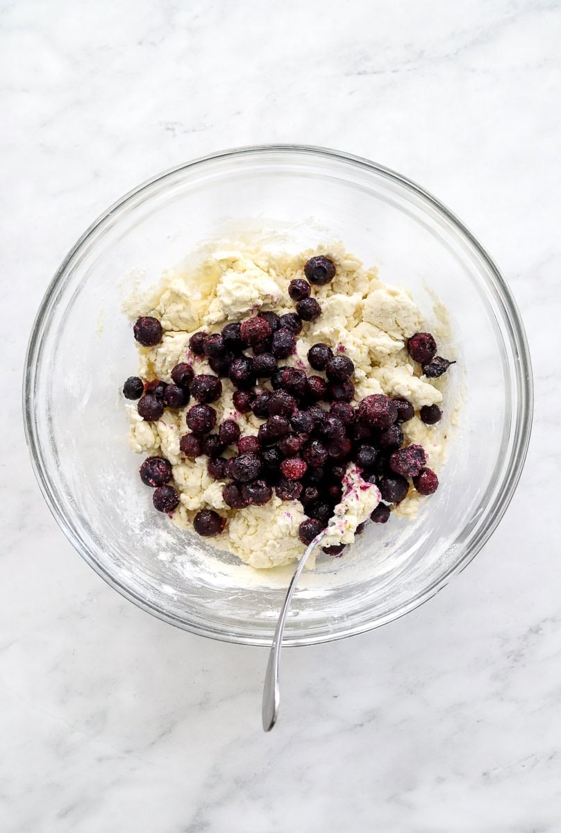 Bagel dough with frozen blueberries added in a bowl with a spoon in the bowl with it.
