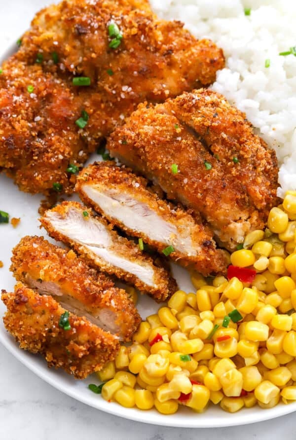Breaded crispy chicken cut into pieces next to corn and fluffy white rice on a white plate.