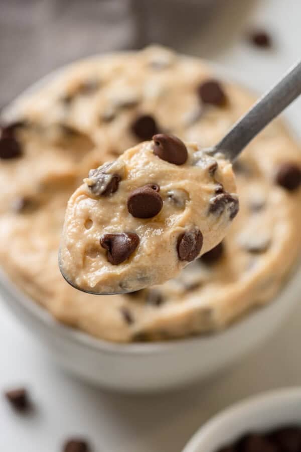 Spoon of cookie dough with more in a bowl behind it with chocolate chips on it.