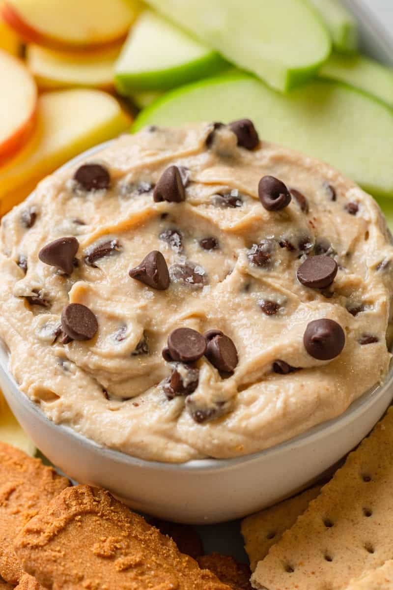 Bowl filled with chocolate chip cookie dough dip with cookies and apple slices around it.