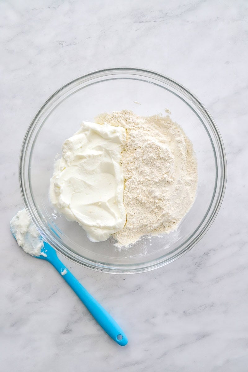 Flour and yogurt in a glass bowl with a blue rubber spatula next to it.